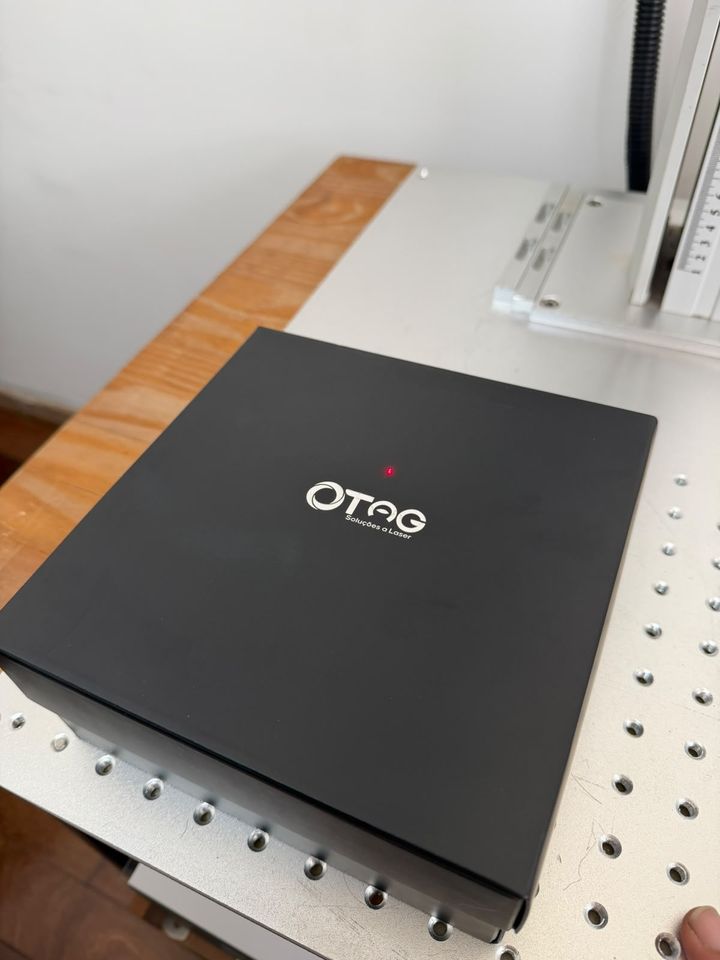 Caderno preto com o logo da TAG gravado, ao lado da máquina